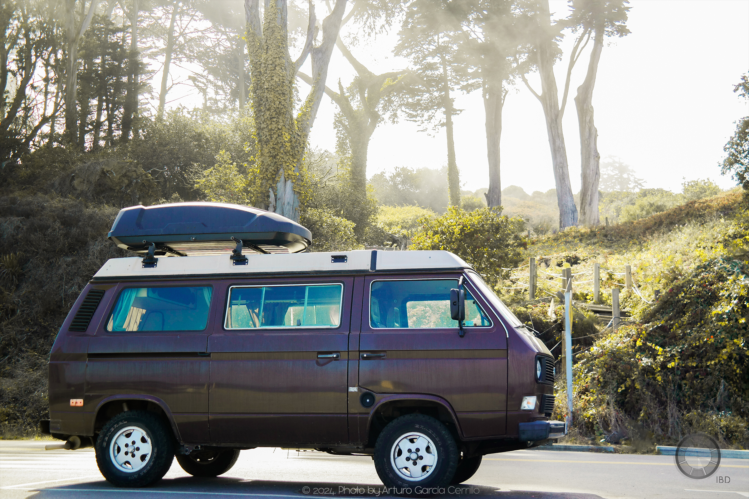 Picture of a brown Old School Volkswagen camping van parked in the street. Behind it is a stairway to a park with tall trees and green plants flooding the background.