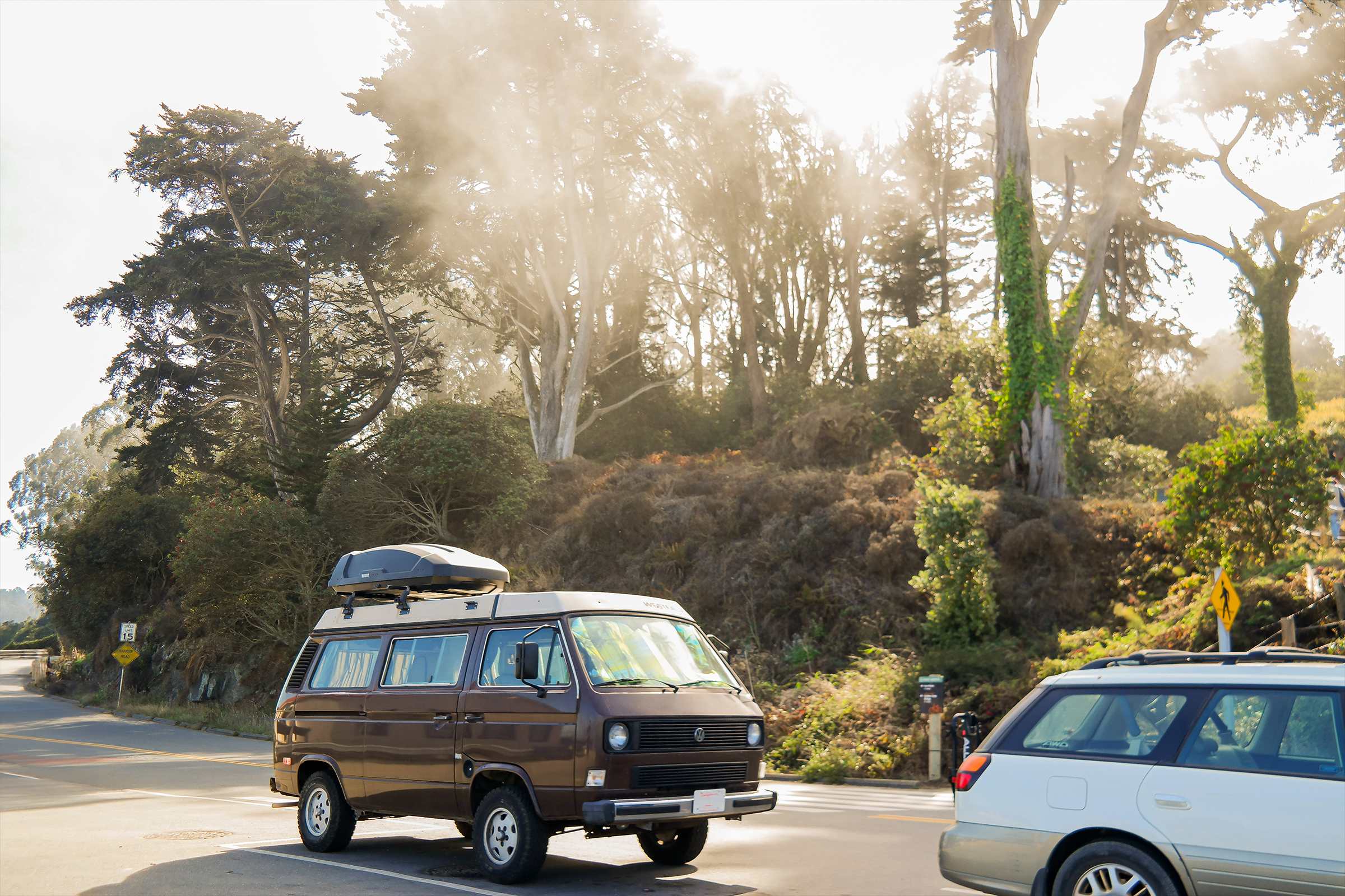 Picture of a brown Old School Volkswagen camping van parked in the street. Woods in the back.